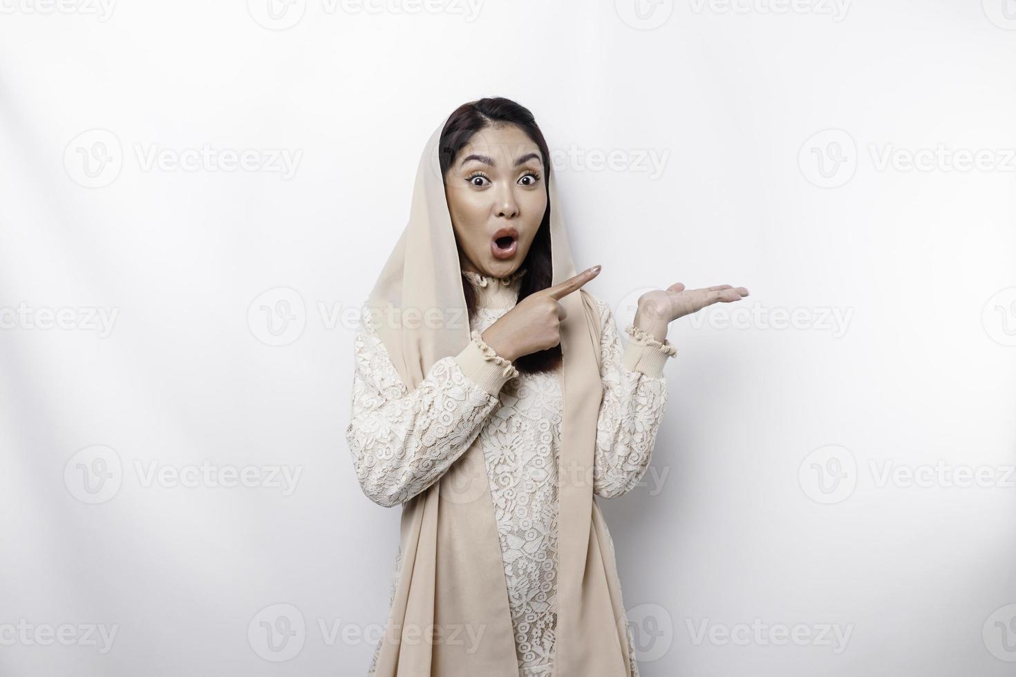 Shocked Asian Muslim woman pointing at the copy space on beside her, isolated by white background photo