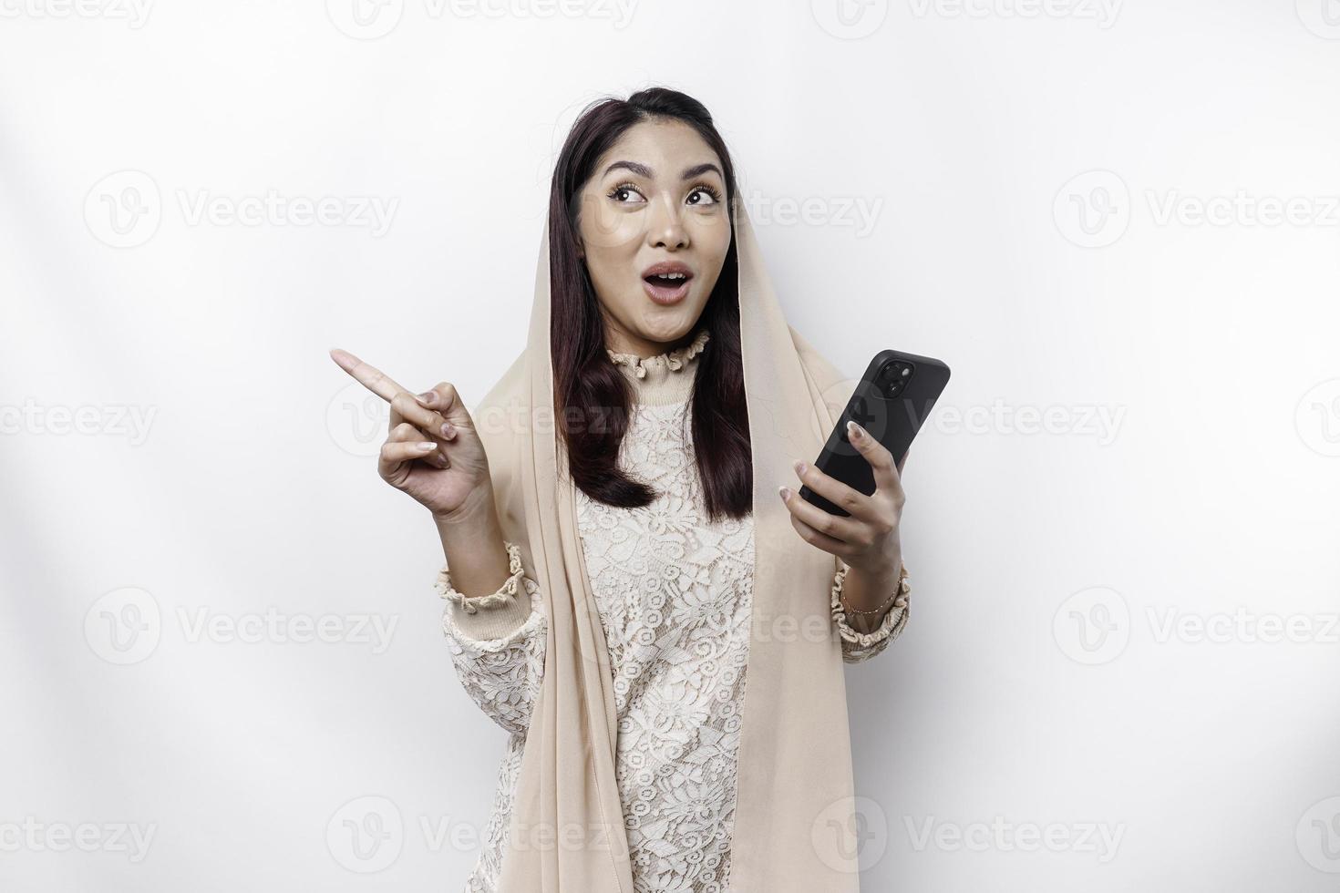 Shocked Asian Muslim woman wearing hijab pointing at the copy space beside her while holding her phone, isolated by white background photo