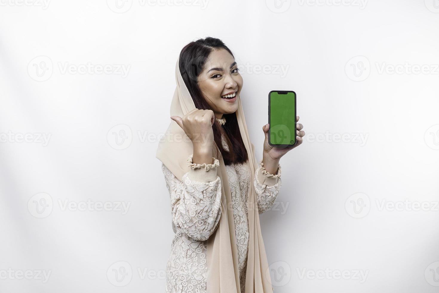 A portrait of a happy Asian Muslim woman wearing a headscarf, showing her phone screen, isolated by white background photo