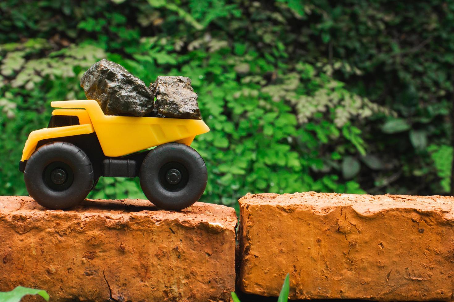 After some edits, a photo of a yellow toy dump truck try to carry pile of stones on a brick.