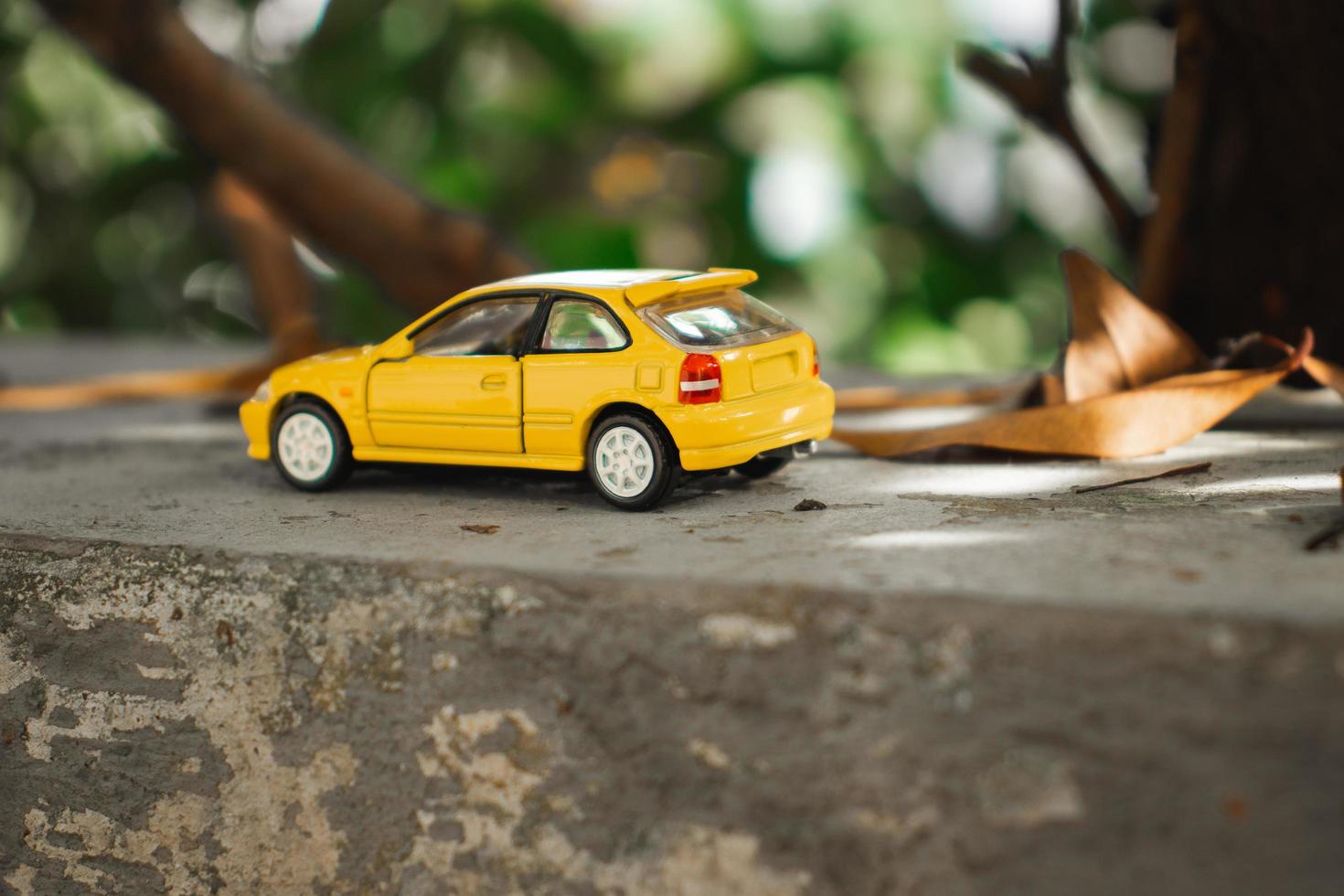 A photo of a yellow toy car placed near a tree, after some edits. Concept for nature adventure.