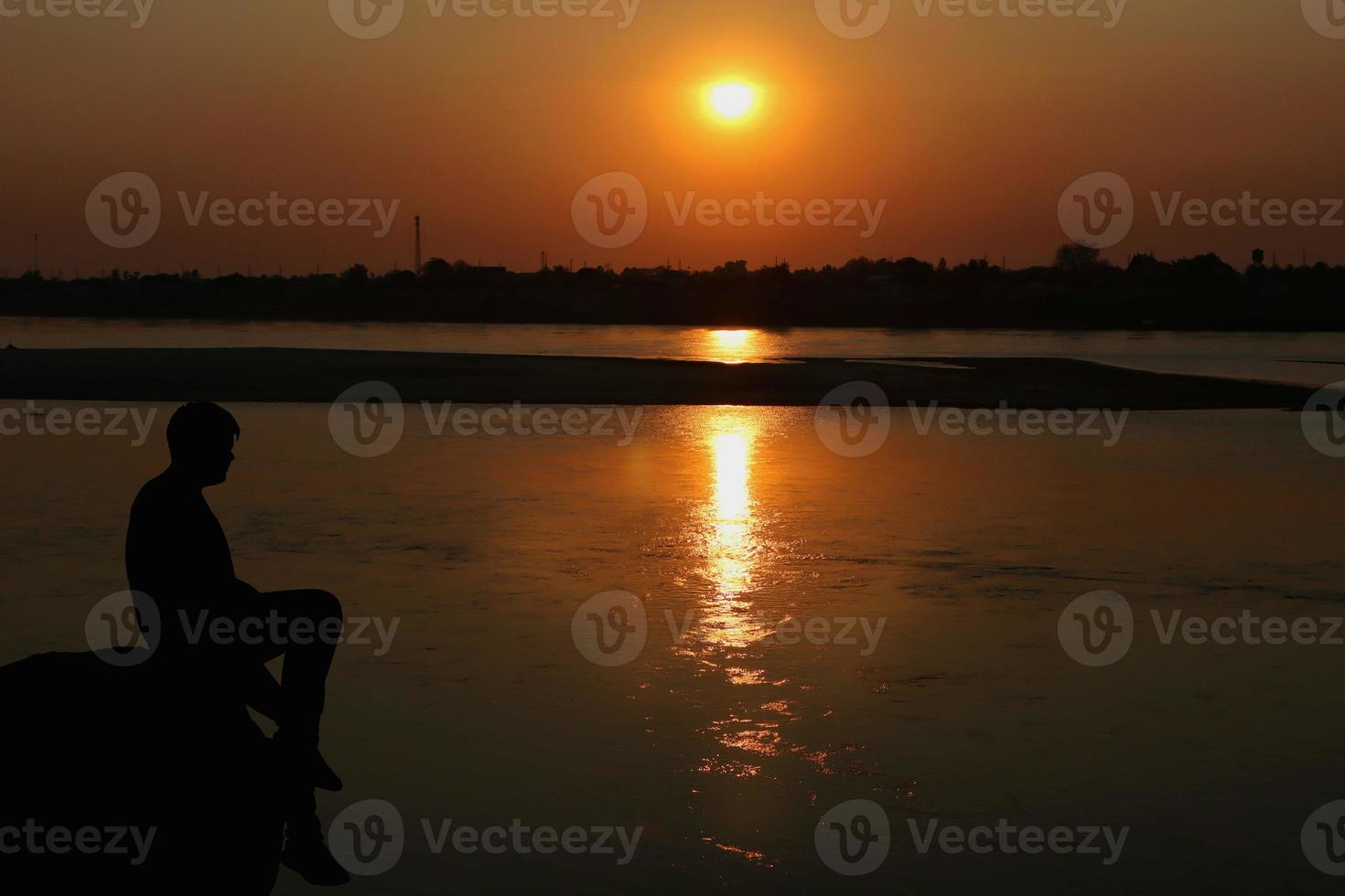 Silhouette of a man sitting on a rock by the river looking at sunset photo