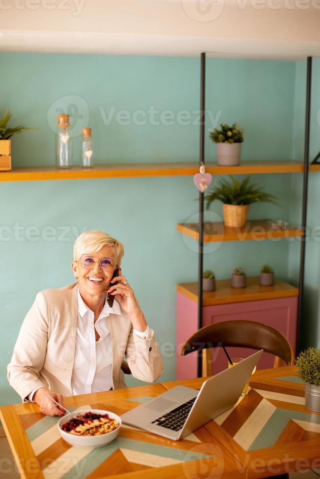 Senior woman using mobile phone while working on laptop and having healthy breakfast in the cafe photo