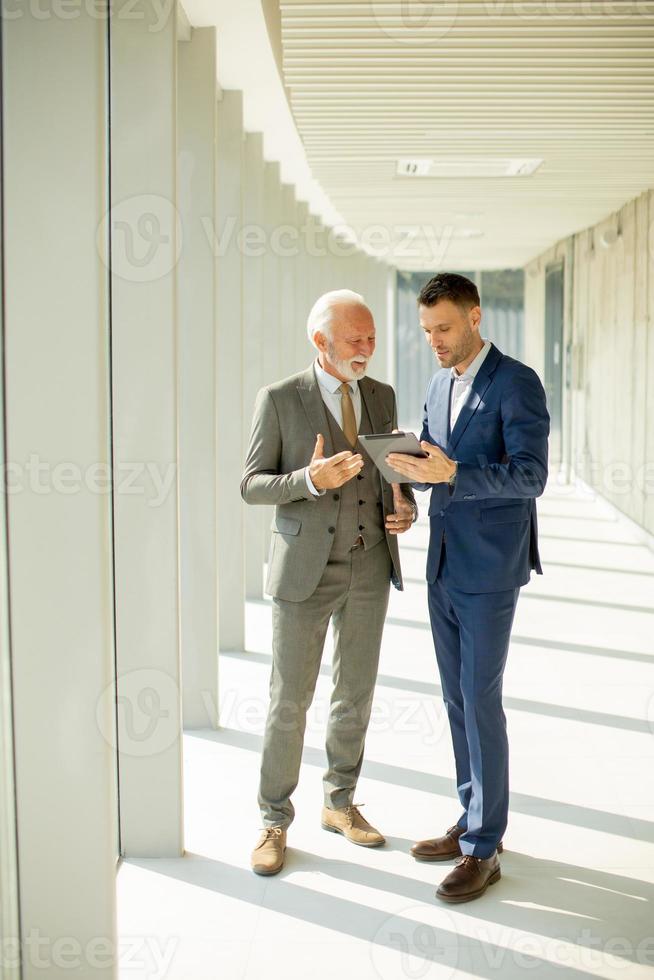 Mature businessman and his young colleague discussing finantial report on   digital tablet in the office corridor photo