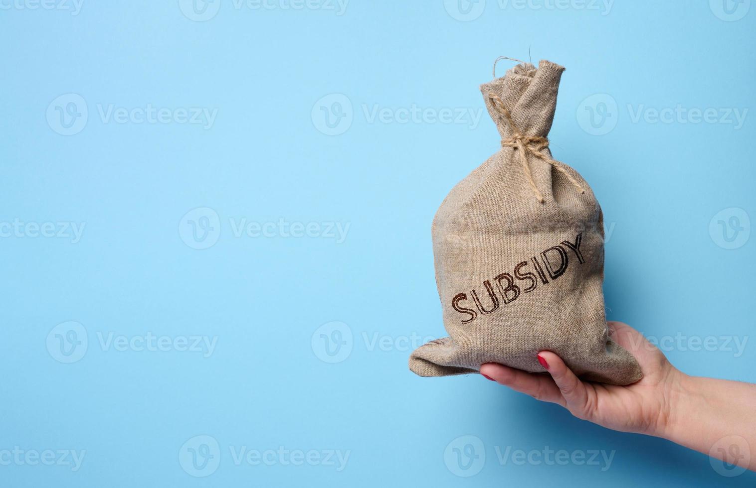 A female hand holds a burlap sack with the inscription subsidy on a blue background, concept of increasing government subsidies to the population photo
