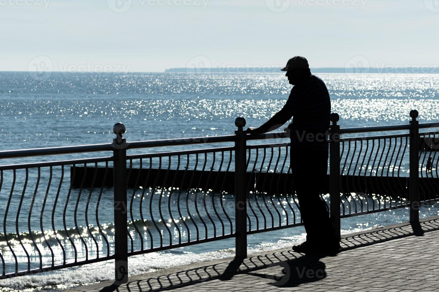 Aged man dreams about life adventures, past traveling and looks at sea. Rear back view, copy space. Old cancer widower misses his wife. Sunny weather and clean blue sea. photo