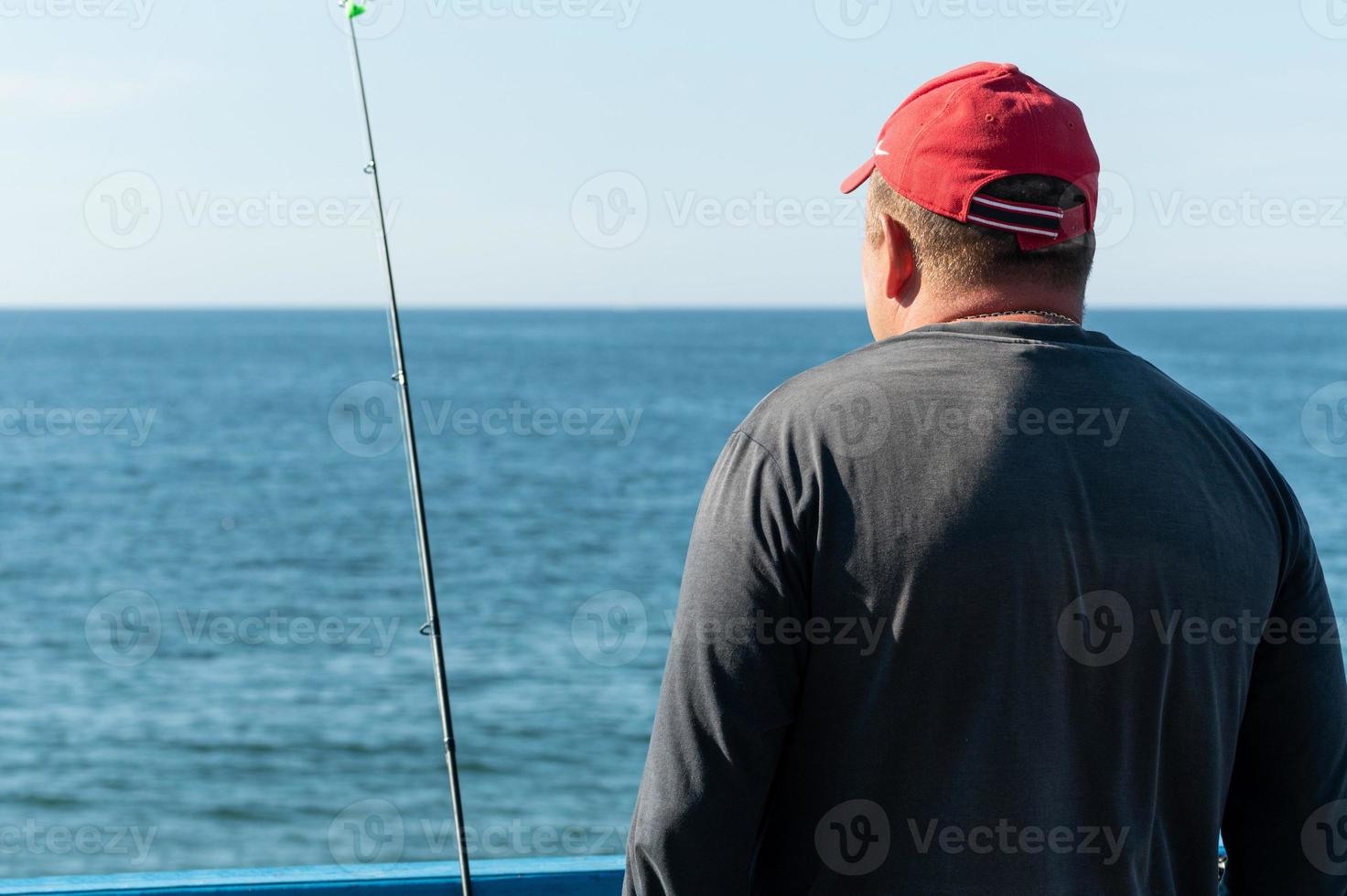 Fisherman fishing from pier. Man with fishing rod in red cap. Windless weather, calm. Copy space. Rich catch, fish place. photo