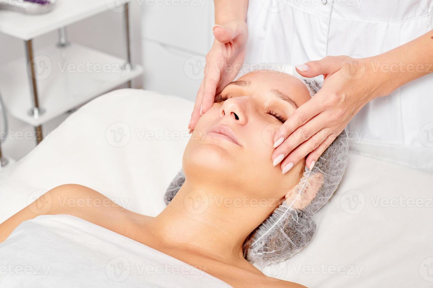 Beautician massages cream mask into woman face skin for rejuvenation, procedure in beauty salon photo