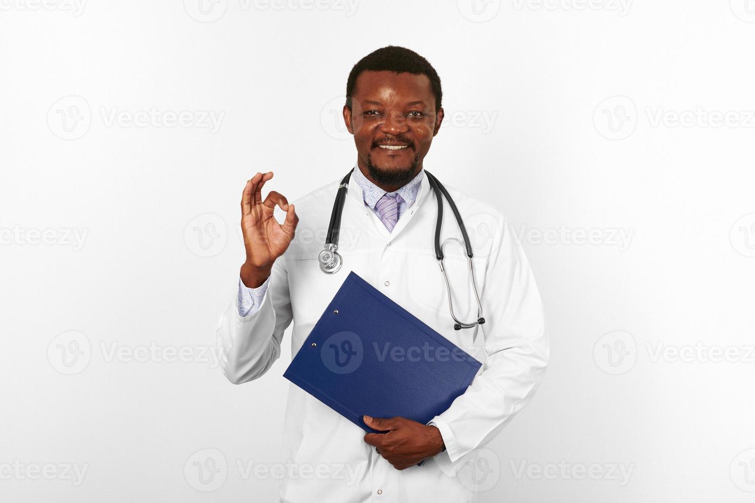 Smiling black bearded doctor man in white robe holds medical clipboard and shows OK gesture photo