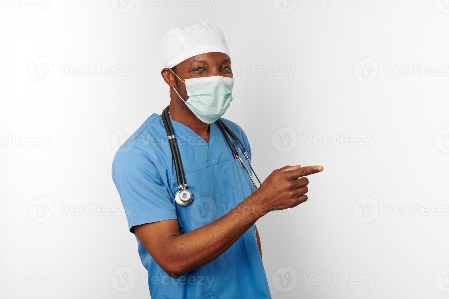 negro cirujano médico hombre en azul Saco blanco gorra y cirujano máscara señalando dedo a Derecha foto