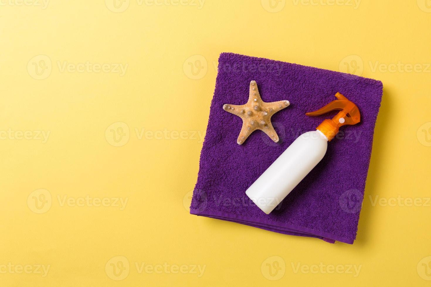 Beach flat lay accessories with copy space. Violet blue and white towel, seashells, staw sunhat and a bottle of sunblock on yellow background. Summer holiday concept photo