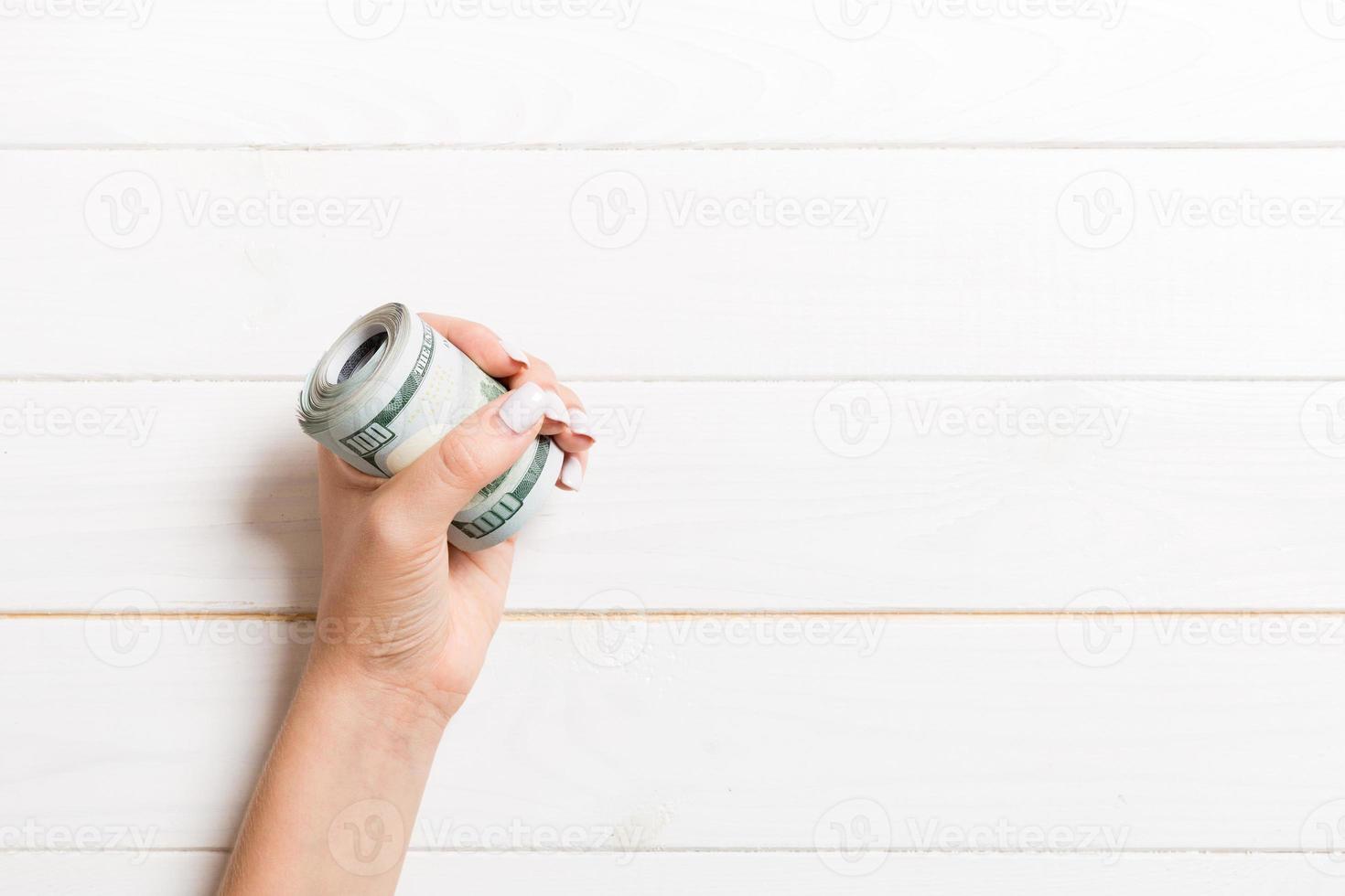 Female hand holding tightly a rolled bundle of money. Top view of one hundred dollars on wooden background. Investment concept with copy space photo