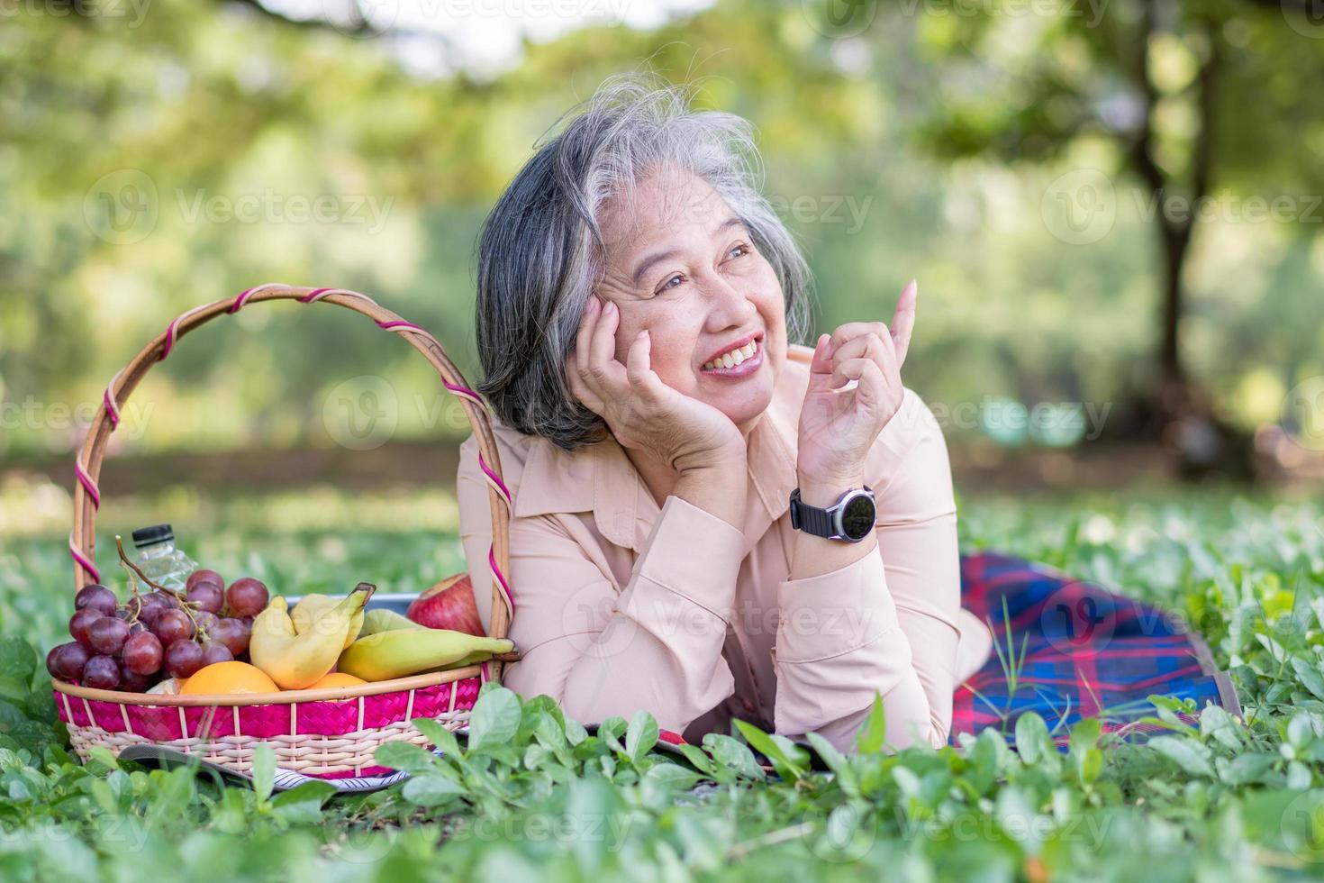 contento asiático antiguo mayor mujer y vestir un salud reloj y acostado en el picnic estera en parque y cesta de Fruta además. concepto de contento mayor mujer después Jubilación y bueno salud foto