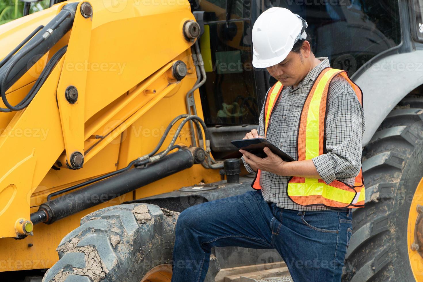 Ingenieria vistiendo un blanco la seguridad casco en pie en frente de el retroexcavadora mirando a hogar construcción trabajo y utilizar el tableta a cheque el Plano con construcción foto