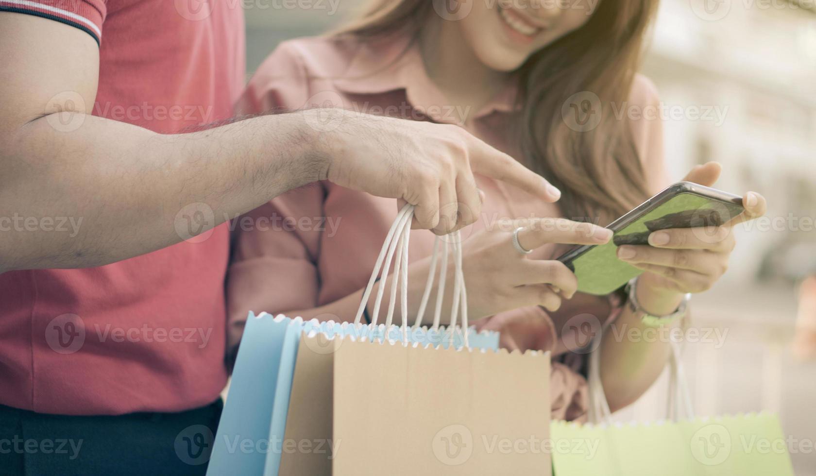 Happy young couple of shoppers walking in the shopping street towards and holding colorful shopping bags in hand and use a smartphone for check promotion. Concept of sale and Black Friday shopping photo