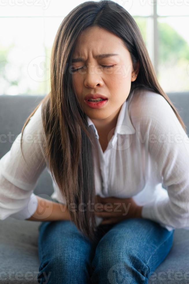 Unhappy Asian woman sitting on the sofa and holding on stomach suffering. Abdominal pain that comes from menstruation, diarrhea, or indigestion. Sickness and healthcare concept photo