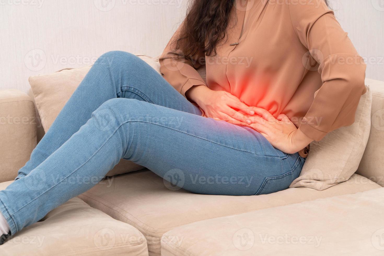 Unhappy Asian woman sitting on the sofa and holding on stomach suffering. Abdominal pain that comes from menstruation, diarrhea, or indigestion. Sickness and healthcare concept photo