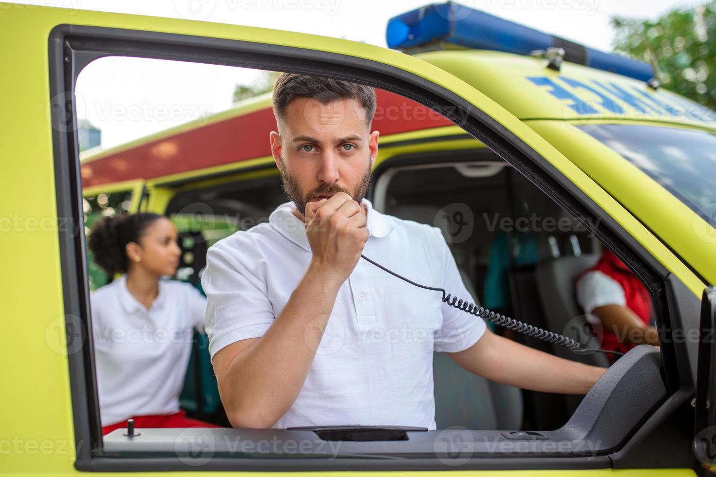 un adulto hermoso masculino paramédico es hablando en un portátil radio mientras sentado en un ambulancia fuera de un clínica. africano americano hembra paramédico en pie por el ambulancia foto