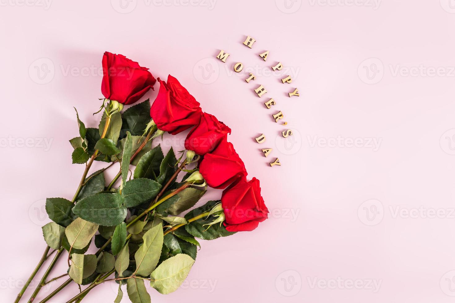 a red bouquet of fresh scarlet roses on a pink background with the text of wooden letters of happy mother's day. top view. flat styling. postcard photo