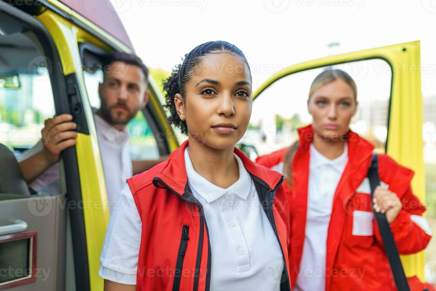 Three multiracial paramedics standing in front of ambulance vehicle, carrying portable equipment photo