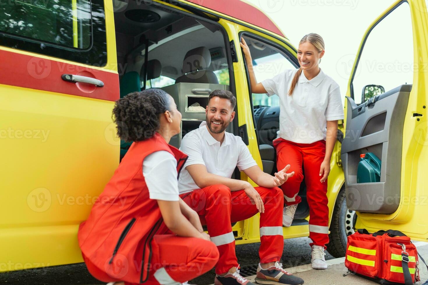 Multi-ethnic group of three paramedics at the rear of an ambulance, climbing in through the open doors. The two women are smiling at the camera, and their male colleague has a serious expression. photo