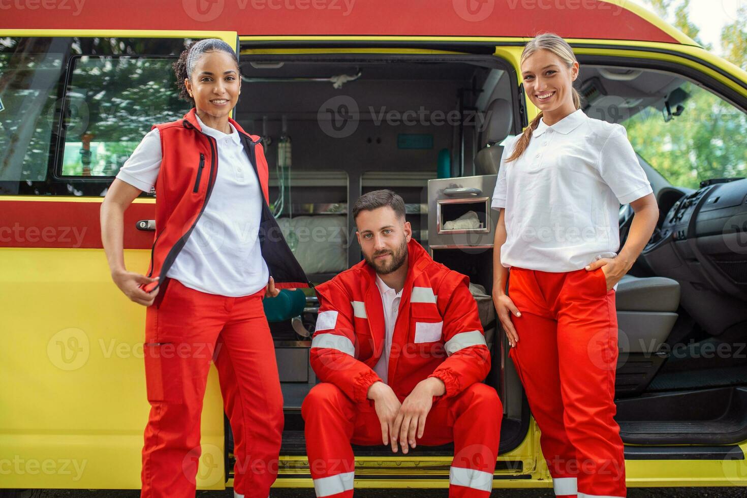 A multi-ethnic group of three paramedics at the rear of an ambulance, climbing in through the open doors. The two women are smiling at the camera, and their male colleague has a serious expression. photo