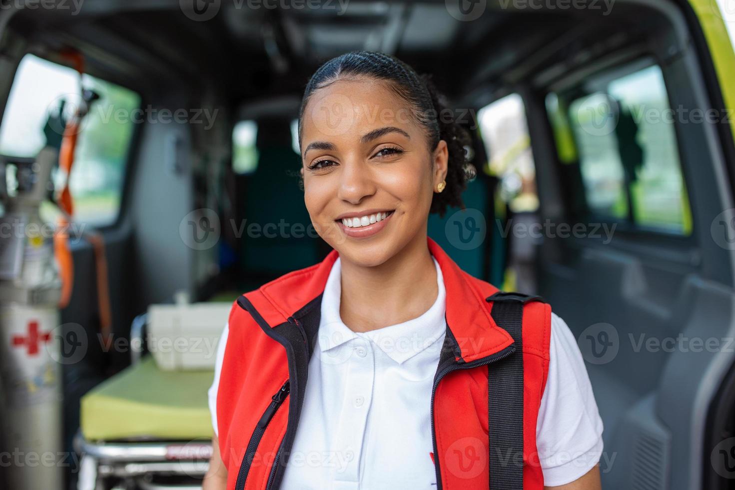Young woman , a paramedic, standing at the rear of an ambulance, by the open doors. She is looking at the camera with a confident expression, smiling, carrying a medical trauma bag on her shoulder. photo