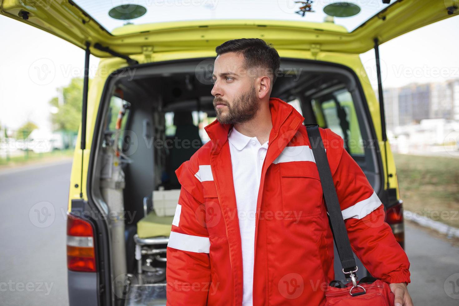 Young man , a paramedic, standing at the rear of an ambulance, by the open doors. He is looking at the camera with a confident expression, smiling, carrying a medical trauma bag on his shoulder. photo