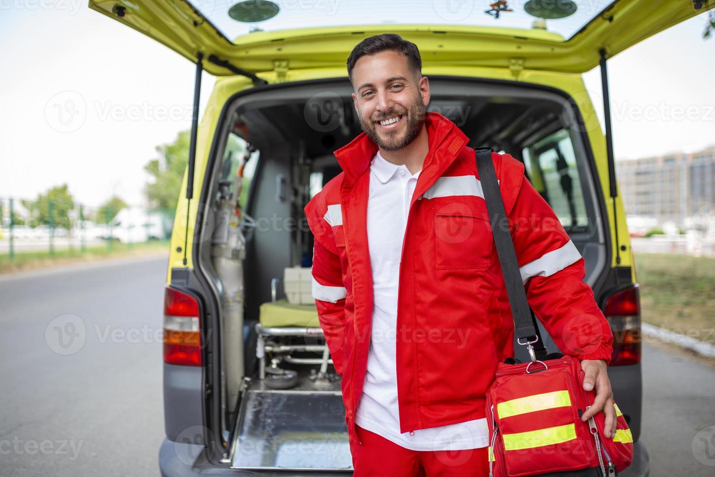 Young man , a paramedic, standing at the rear of an ambulance, by the open doors. He is looking at the camera with a confident expression, smiling, carrying a medical trauma bag on his shoulder. photo
