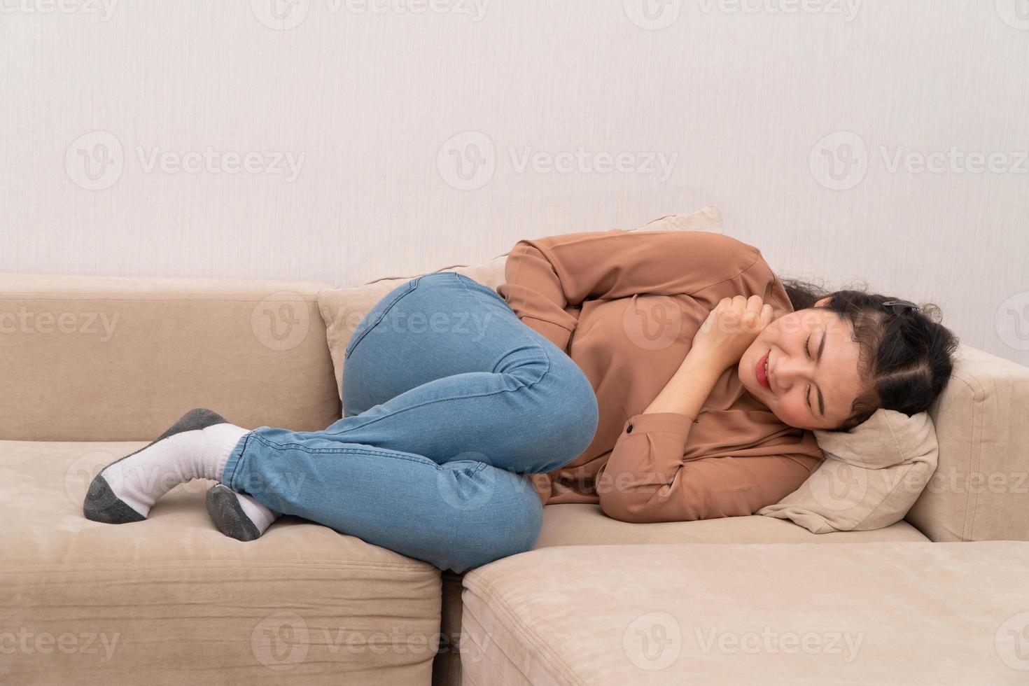 Unhappy Asian woman sitting on the sofa and holding on stomach suffering. Abdominal pain that comes from menstruation, diarrhea, or indigestion. Sickness and healthcare concept photo