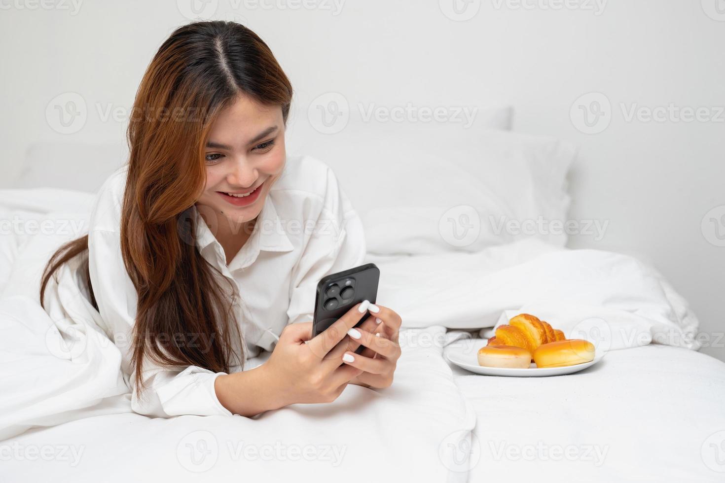 Woman in white nightgown waking up on weekend morning resting and relaxing playing with laptop mobile phone Eating bread and drinking tea in glass inside white bedroom.  Morning vacation concept. photo