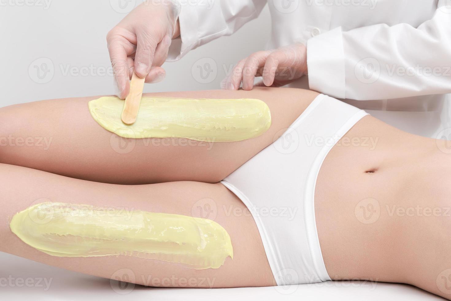 Beautician in gloves applying green hot wax on slim woman legs using spatula while woman lying photo