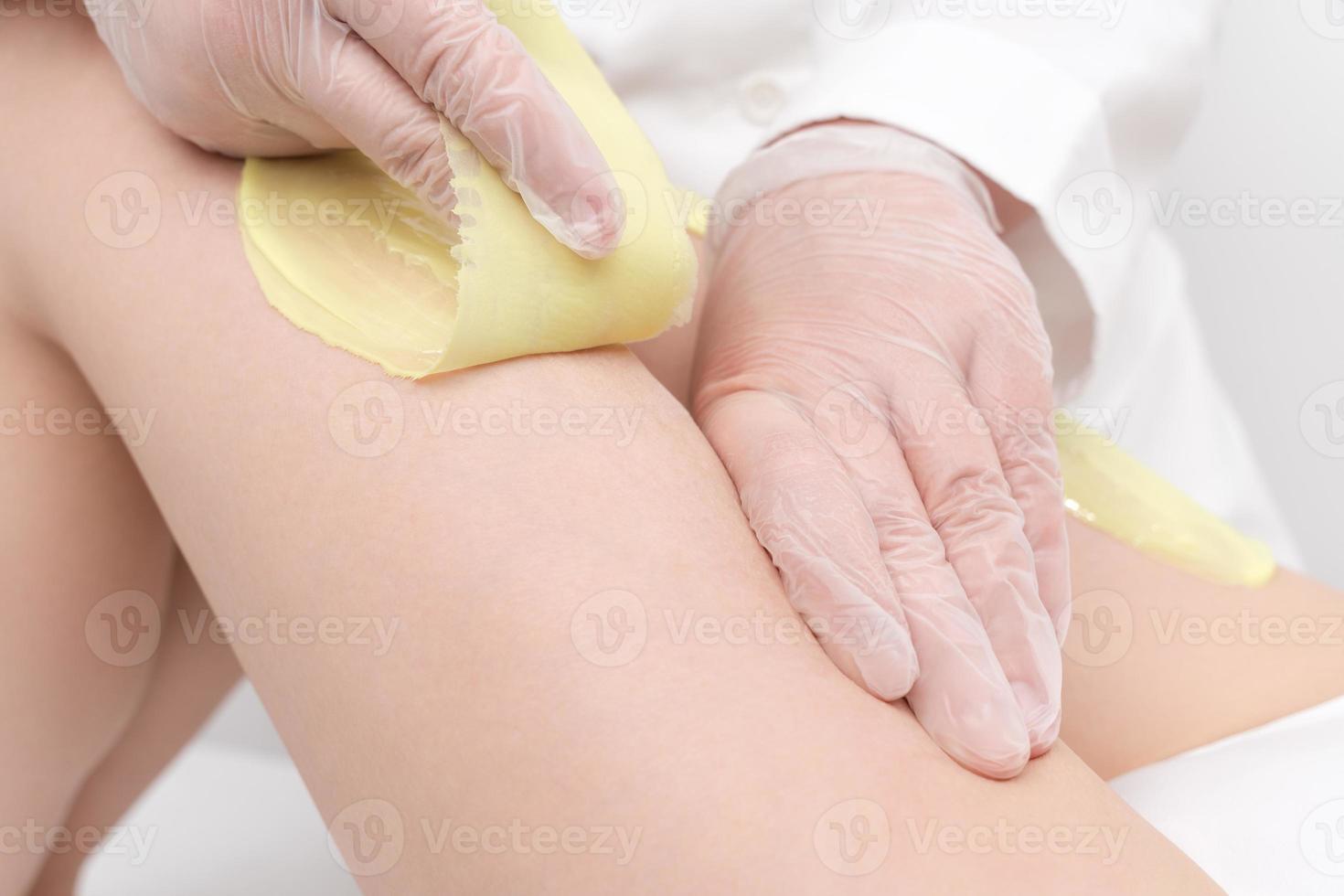Closeup of cosmetologist hands doing depilation to woman legs with green hot wax in beauty salon photo
