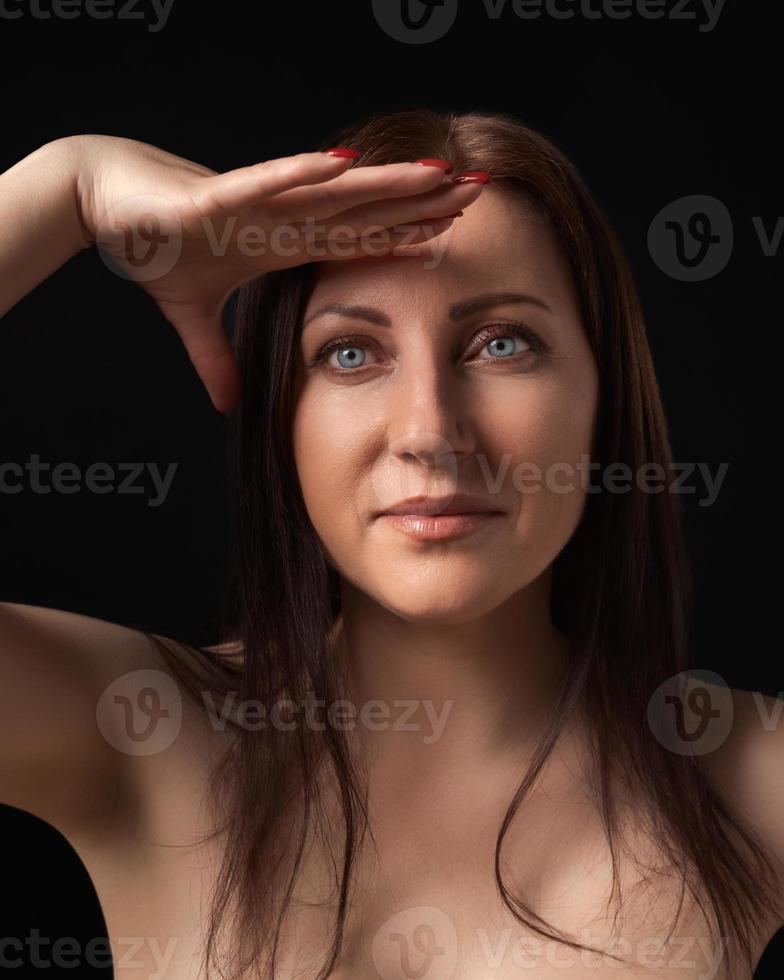 Pretty brunette woman gracefully raising hand to forehead and looking at camera on black background photo