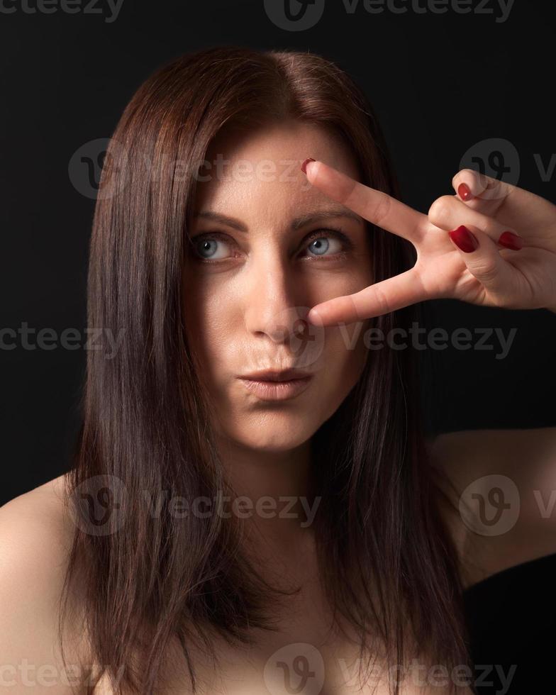 Studio portrait of woman showing victory or peace gesture with fingers near eye on black background photo