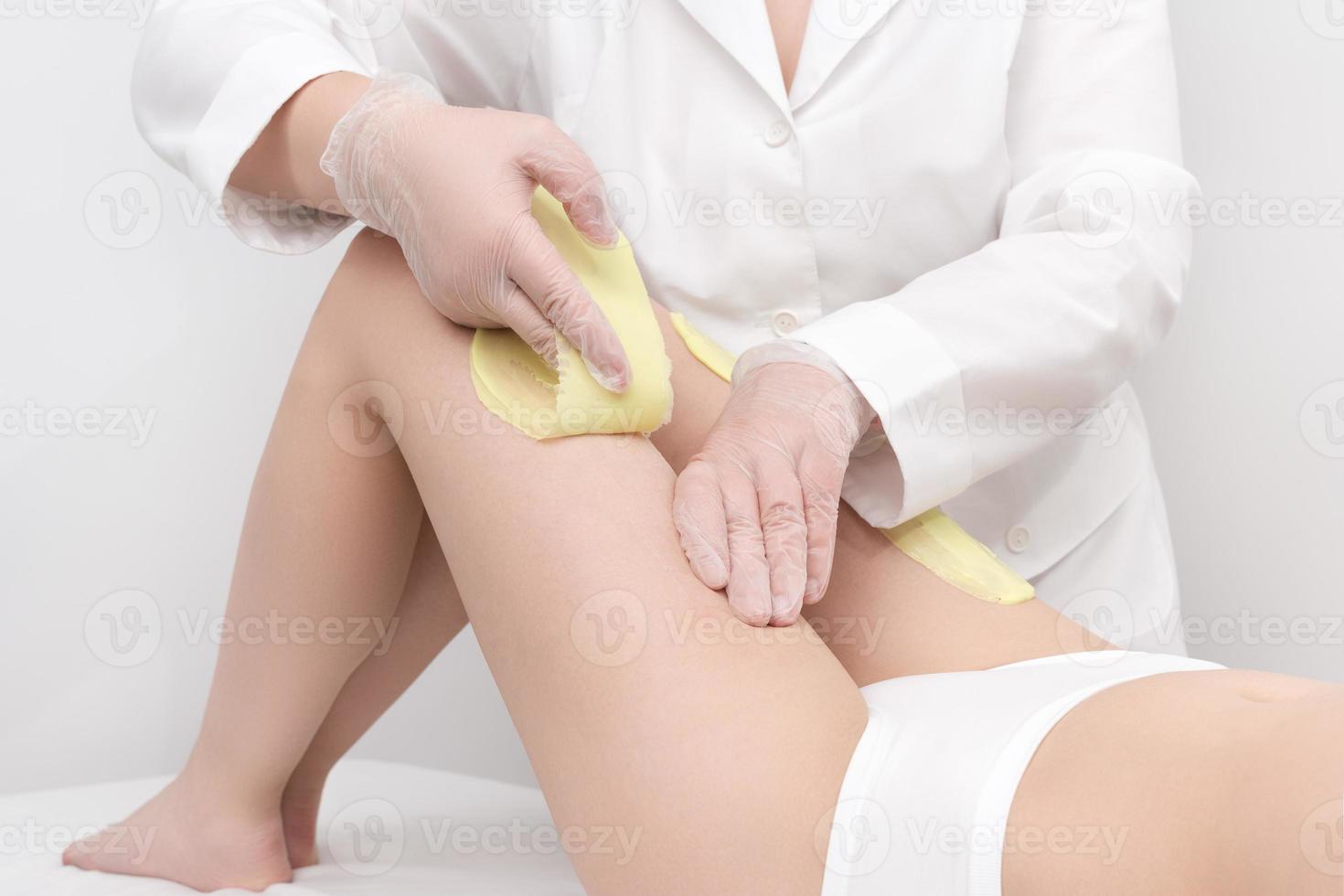 Unrecognizable beautician in white coat removal hair on women leg. Depilation procedure with hot wax photo