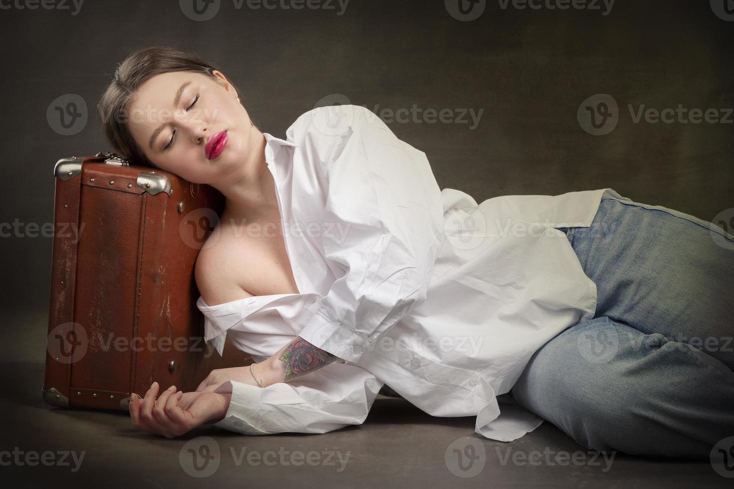 A beautiful girl with closed eyes lies on an old suitcase on a gray background. photo