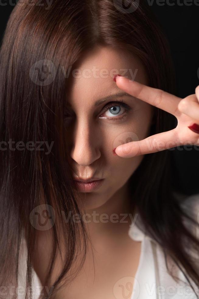 retrato de grave mujer participación dos dedos cerca ojo, demostración victoria gesto, mirando a cámara foto