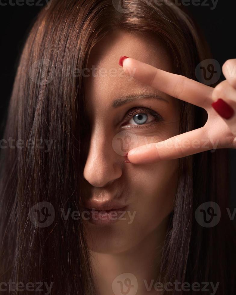 Closeup portrait woman showing victory or peace gesture with fingers near eye and looking at camera photo