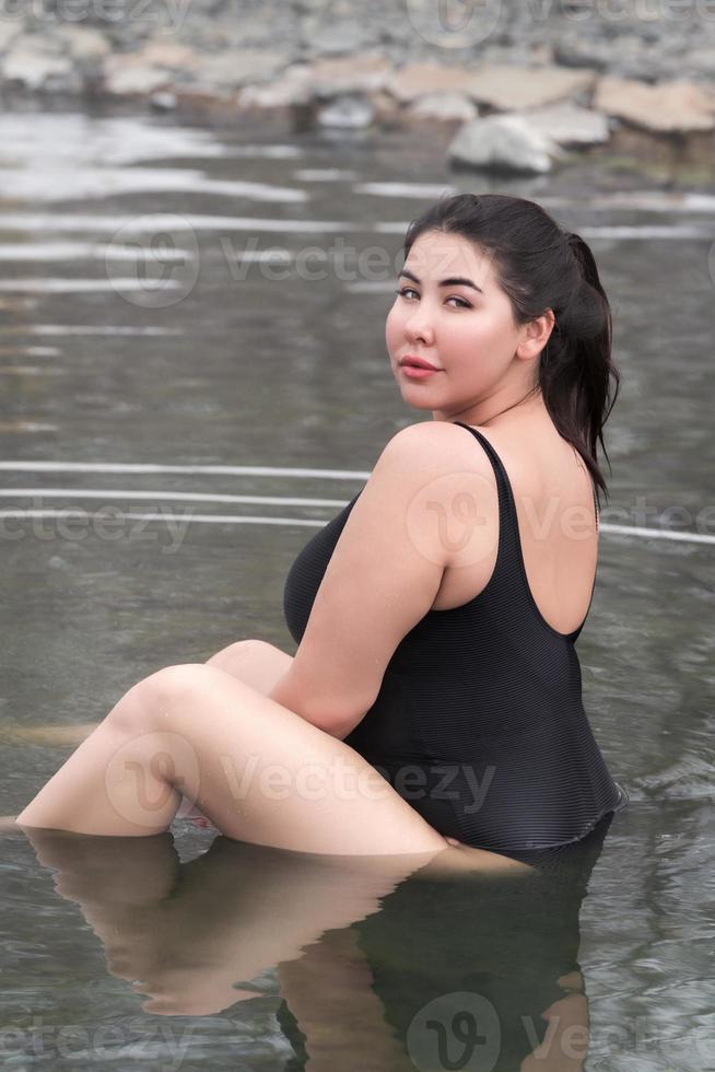 Overweight young woman in black swimsuit sitting in outdoors pool at spa, looking over shoulder photo