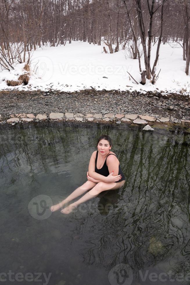 grande Talla joven modelo en negro baños traje de baño sentado en al aire libre piscina a spa, invierno endurecimiento foto