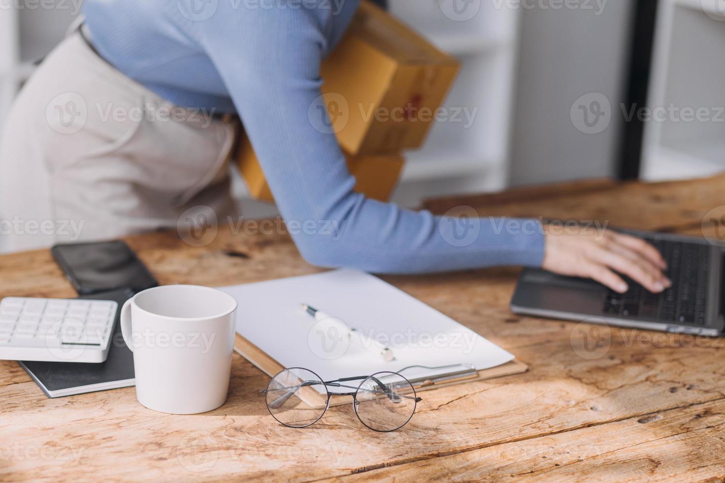 Startup SME small business entrepreneur of freelance Asian woman wearing apron using laptop and box to receive and review orders online to prepare to pack sell to customers, online sme business ideas. photo