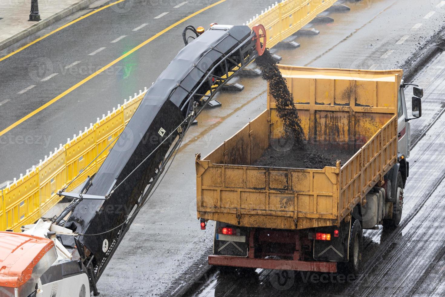 Milling machine working and removing asphalt from the road and pouring it into a truck. Road construction or rehabilitation on a rainy day photo