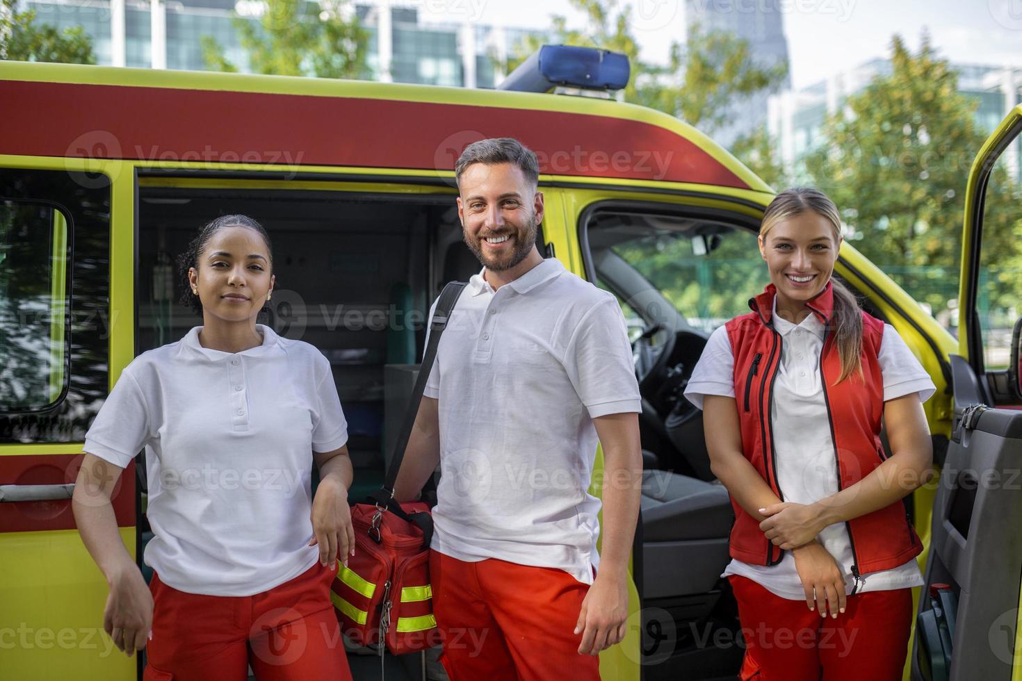 Three multiracial paramedics standing in front of ambulance vehicle, carrying portable equipment photo