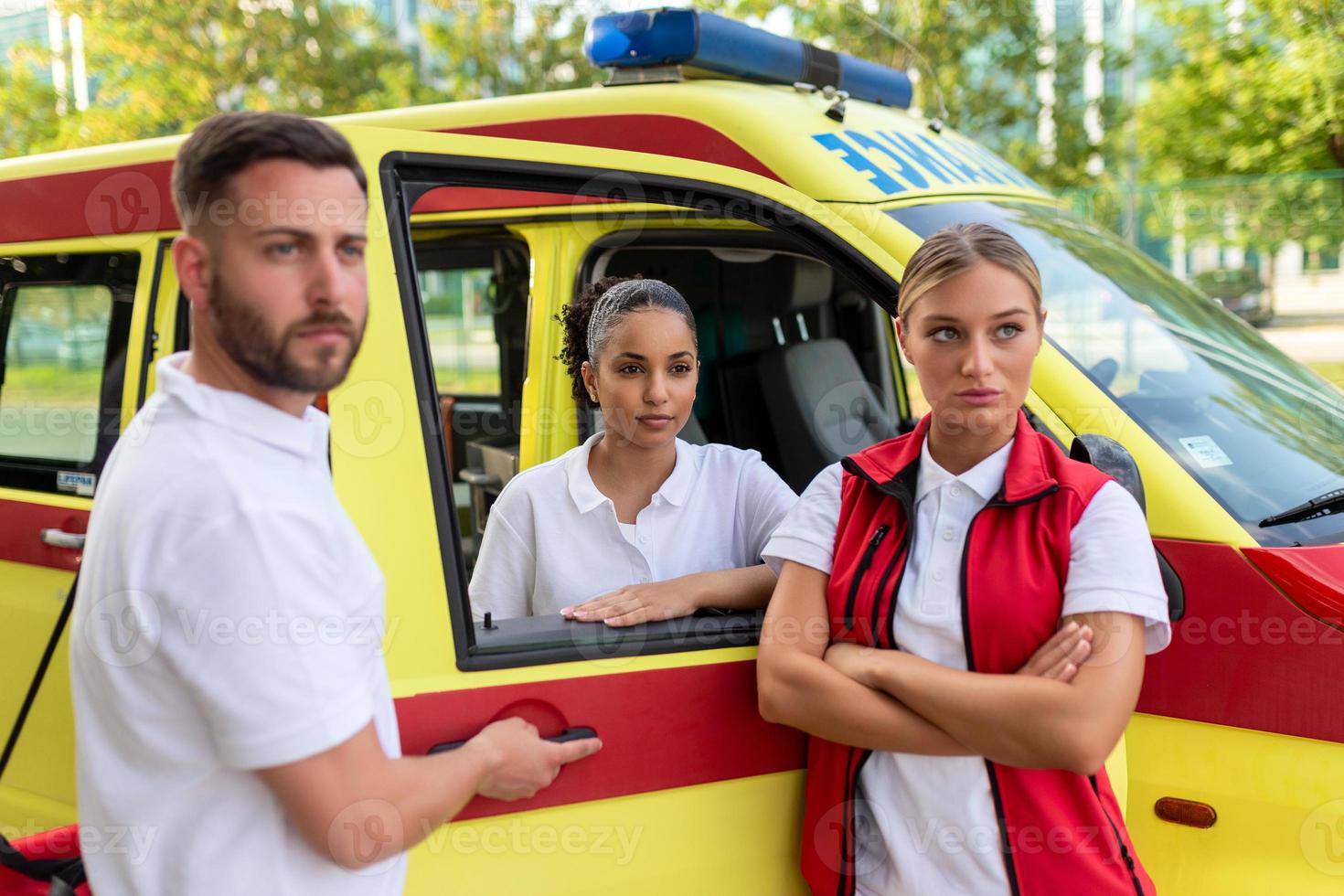 Tres paramédicos en pie por el ambulancia, hablando. masculino paramédico que lleva un médico trauma bolsa. grupo de Tres paramédicos en pie en frente de ambulancia con sonrisa. foto