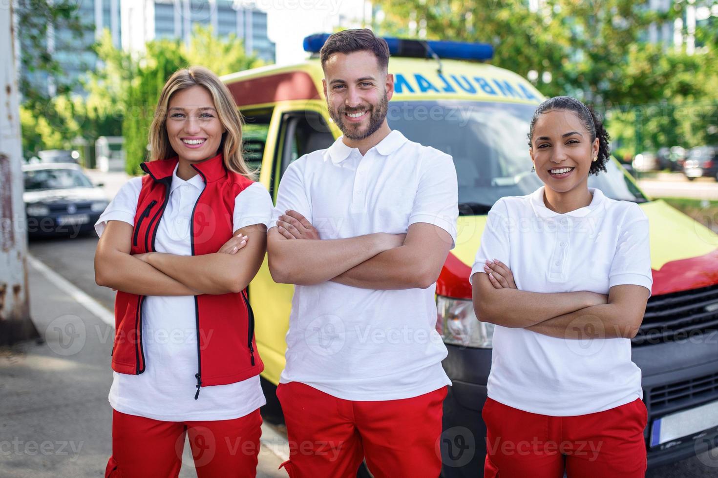 multiétnico grupo de paramédicos en pie a el lado de un ambulancia con abierto puertas su compañero de trabajo que lleva un médico trauma bolsa. ellos son sonriente a el cámara. foto