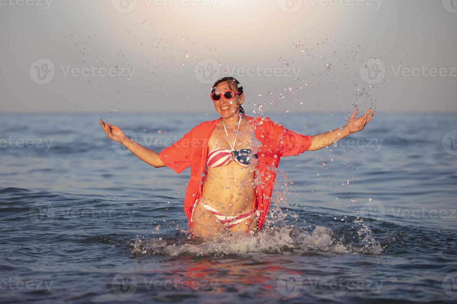 Happy middle-aged woman stands in sea water with raised arms in a swimsuit with an American flag print. photo