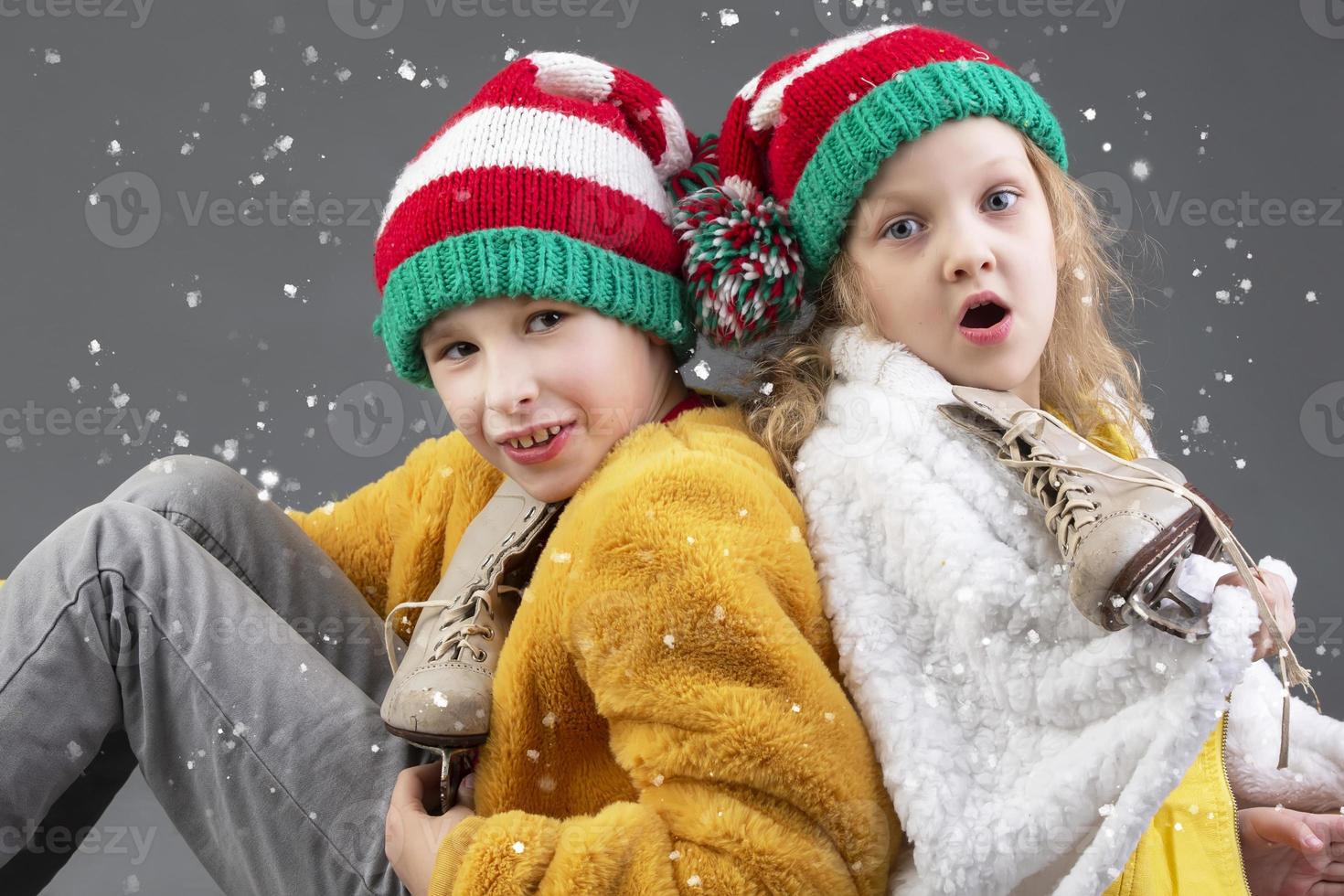 Funny little girl and boy in knitted Christmas hats and vintage ice skates sit with their backs to each other and look at the falling snow on a gray background. Happy Christmas kids. photo