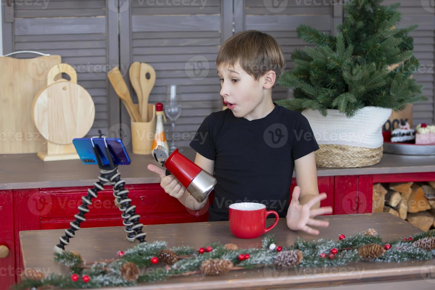 A portrait of a confident and creative child blogger talking to the camera at home in the kitchen, he is vlogging. A little boy is recording a stream on a smartphone. photo