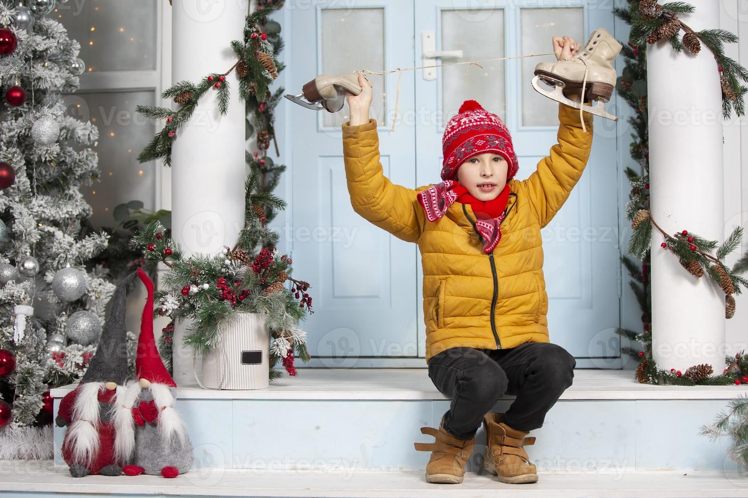 hermoso pequeño chico en Navidad. gracioso niño sostiene antiguo patines y se sienta en el porche de un casa decorado con Navidad árbol decoraciones foto